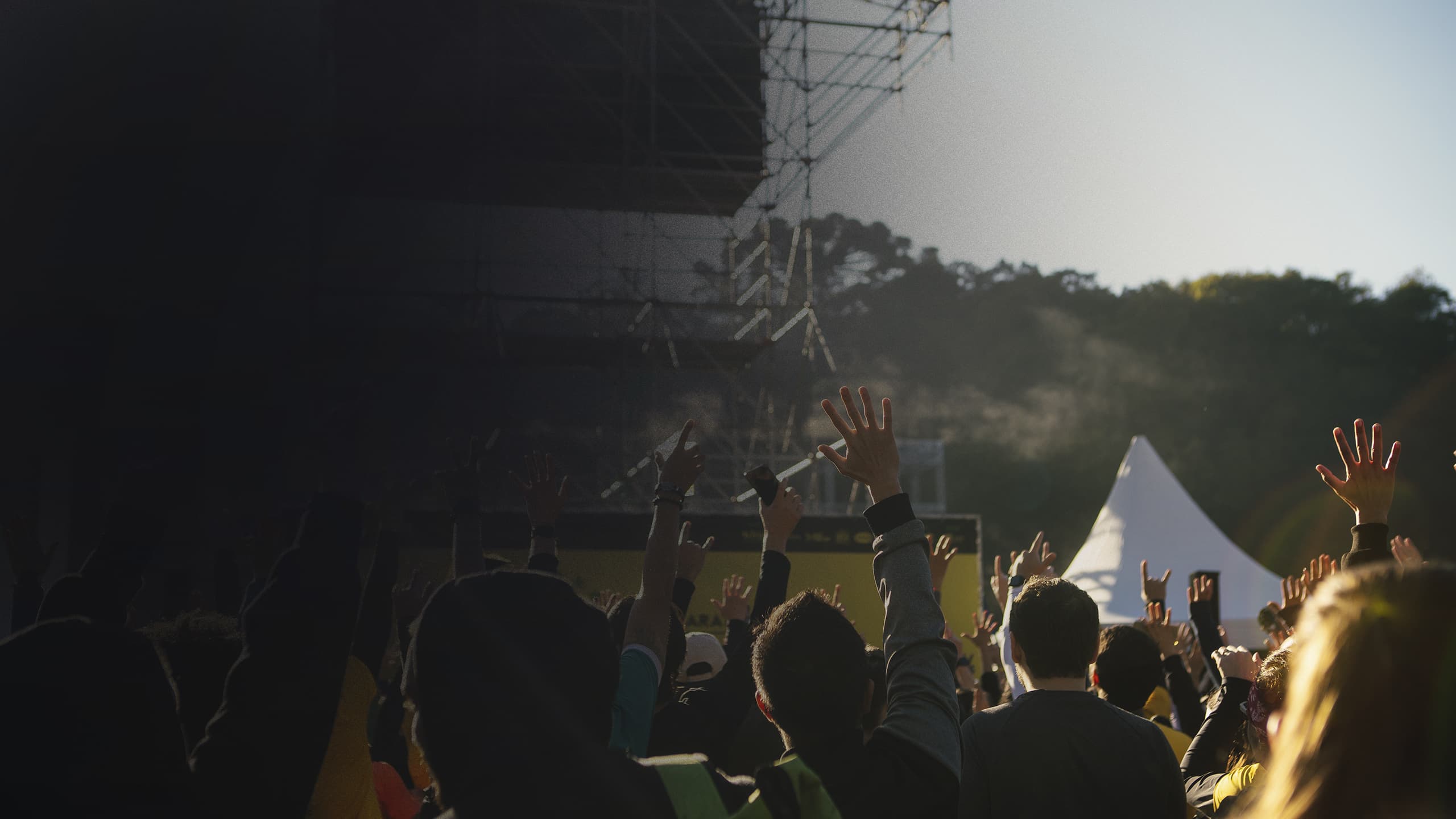 Público de mãos erguidas em frente ao palco, no fim da tarde
